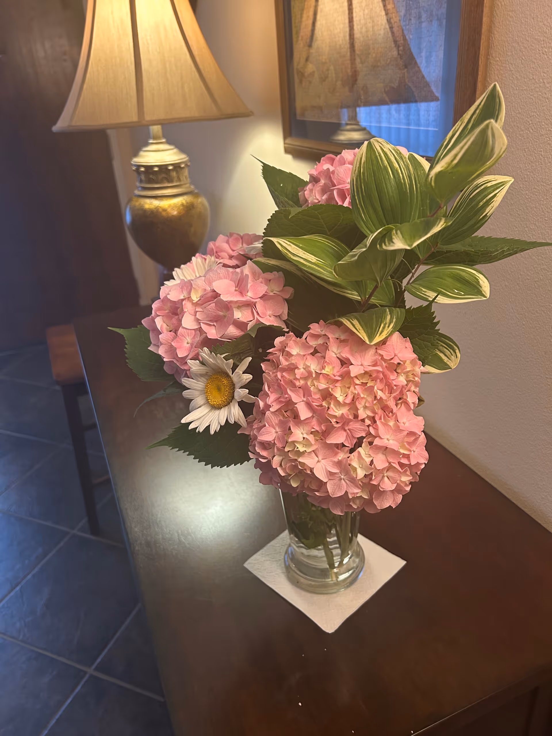 Vase of pink hydrangeas and greenery on a dark wooden table beside a lamp.