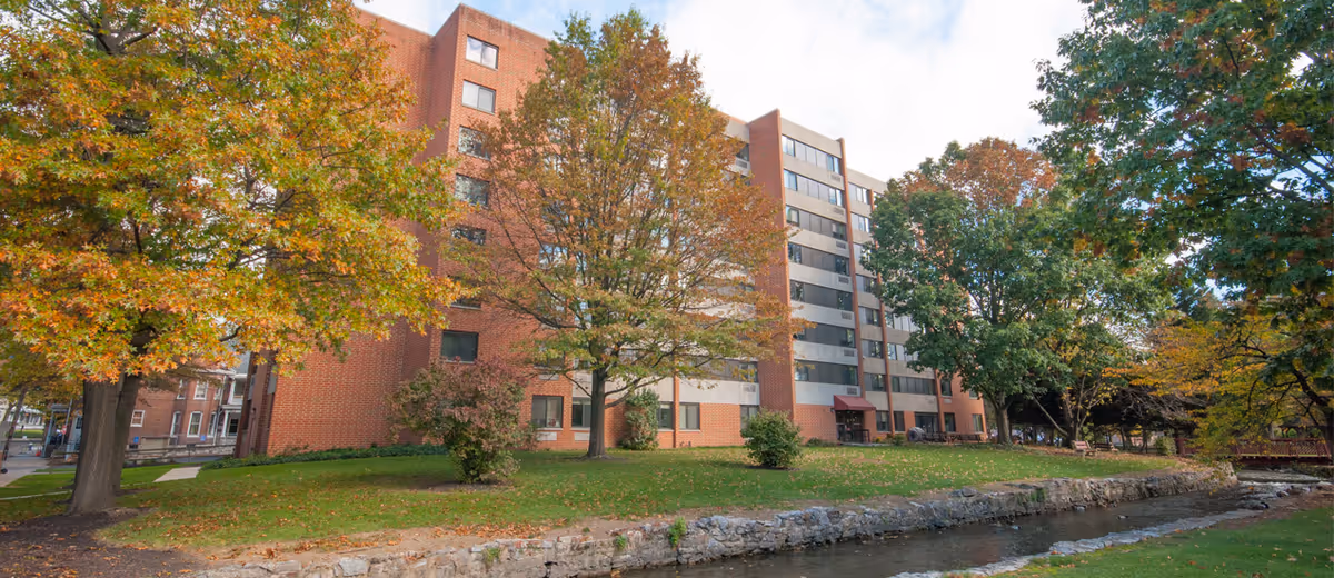 Brick senior living building with trees, riverfront lawn, and autumn foliage.