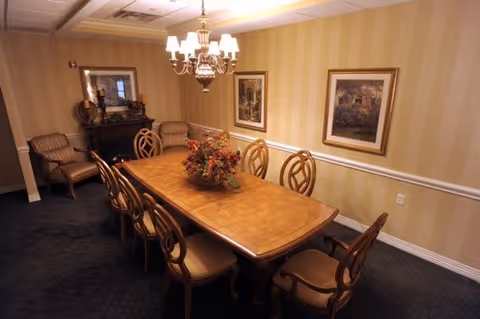 A formal dining room with a long wooden table surrounded by eight matching chairs. A floral centerpiece decorates the table. The room has striped beige wallpaper, two framed paintings on the wall, a chandelier overhead, and a fireplace with a mirror above it. Two armchairs are placed near the fireplace.