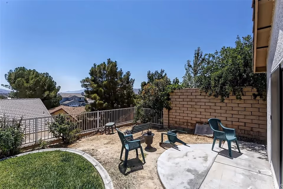 Outdoor patio area with three green plastic chairs, one of which is tipped over, a small round fire pit, and a concrete and dirt ground surrounded by a low brick wall and metal railing. Trees and houses are visible in the background under a clear blue sky.