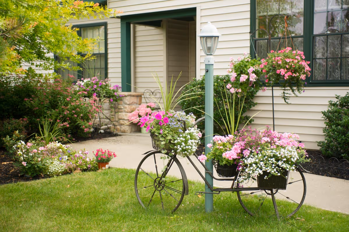 Front of a retirement community building with a decorative bicycle planter, lamppost, and colorful flower beds.