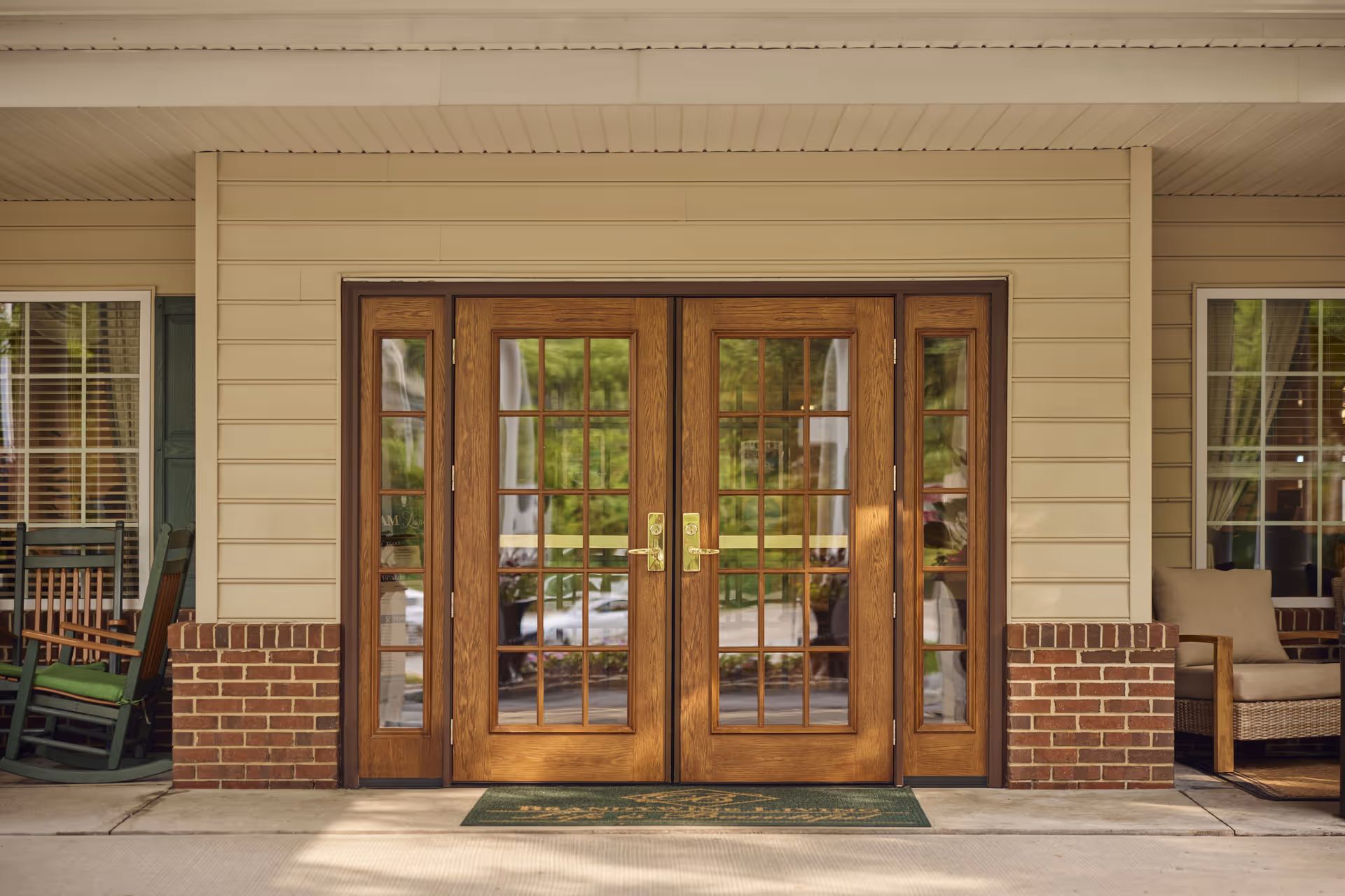 Entrance of Brandywine Middlebrook Crossing by Monarch featuring double wooden doors with glass panes, flanked by windows with white blinds. There are green rocking chairs with cushions on the left side and a cushioned wicker chair on the right side under a covered porch.