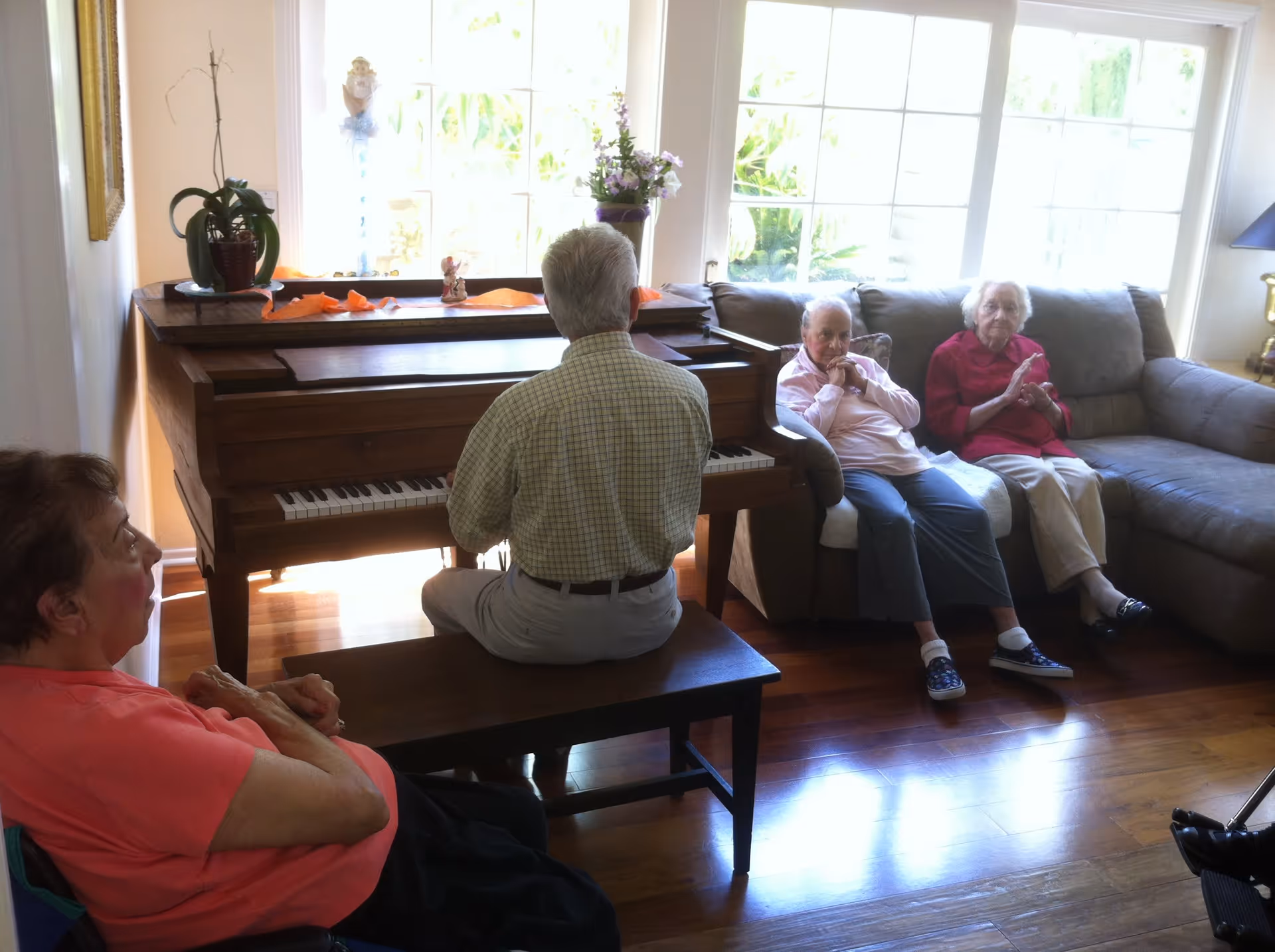 An elderly man playing a wooden piano in a living room with large windows letting in natural light. Three elderly women are seated nearby, two on a couch and one in a chair, listening attentively. The room has wooden floors and is decorated with plants and small ornaments on the piano.