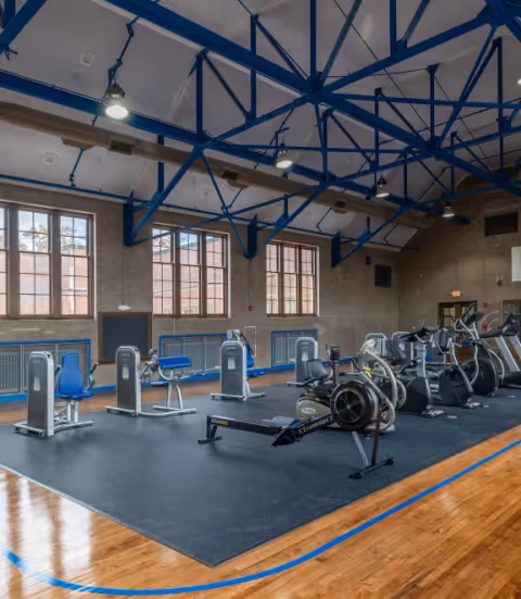 Spacious indoor gym with exercise machines and a rowing machine under blue-painted exposed ceiling beams and large windows.
