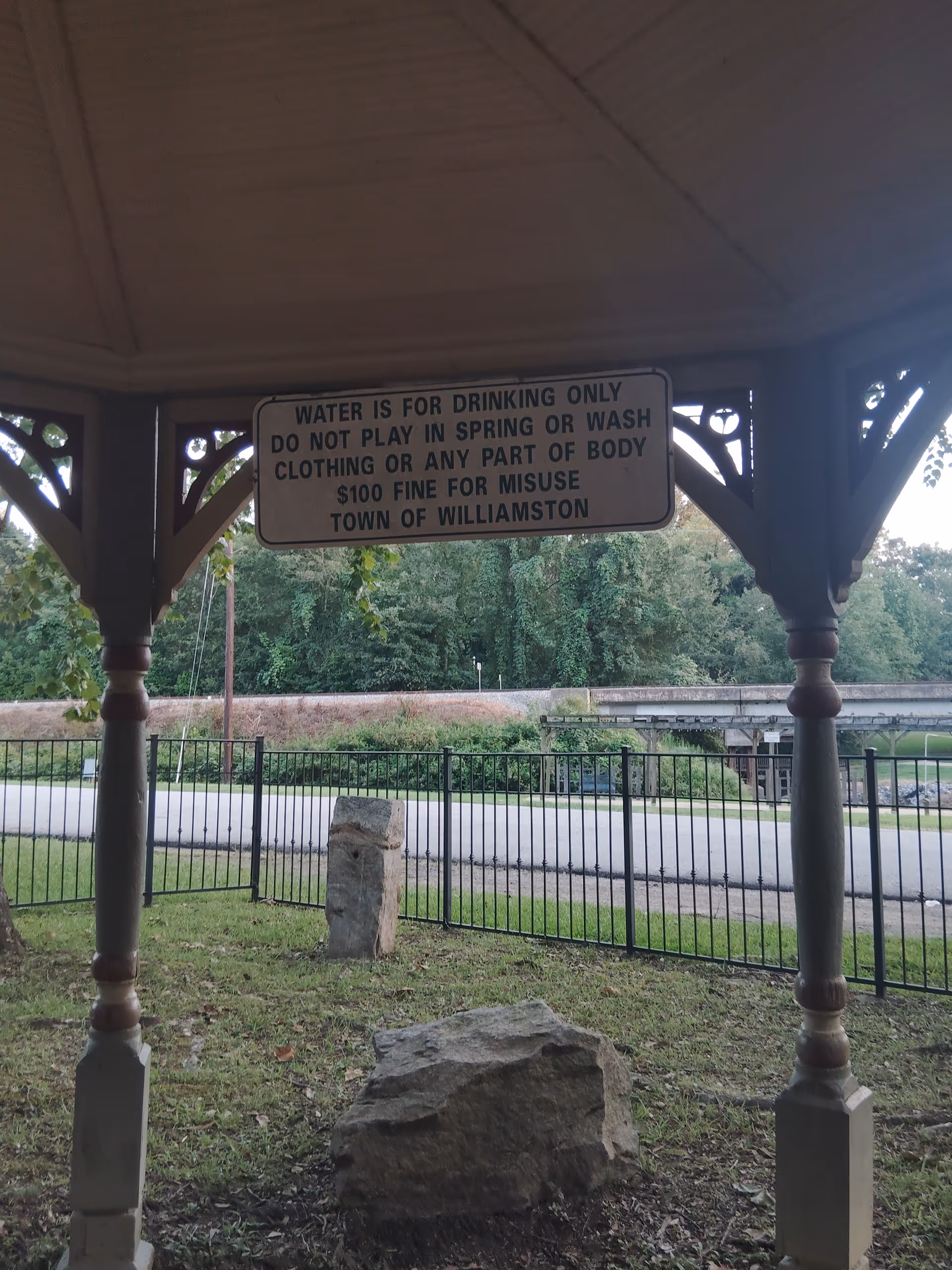 An outdoor gazebo features a sign about drinking water, with surrounding rocks and a fence.
