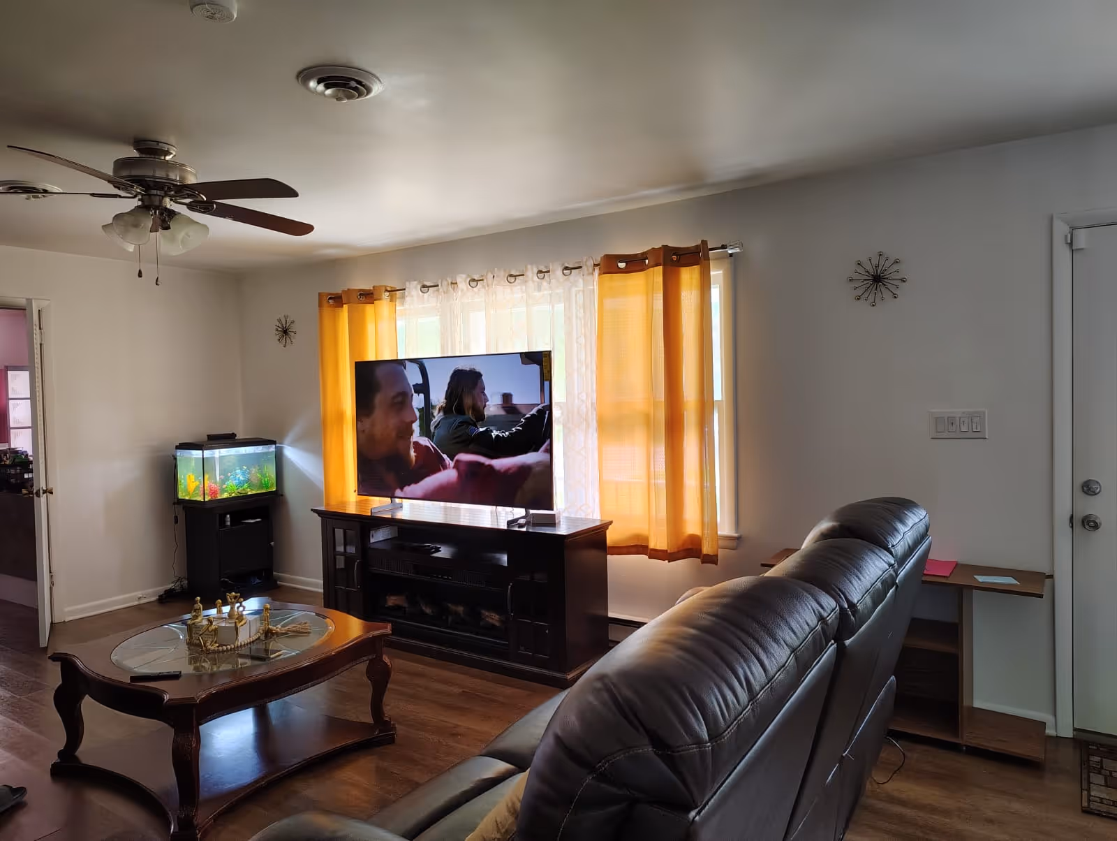 Living room with brown leather sofa, TV on stand, coffee table, and orange-curtained windows.
