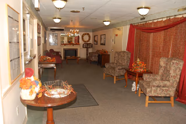A long furnished common lounge with armchairs, side tables, autumn decorations, and a fireplace at the far end.