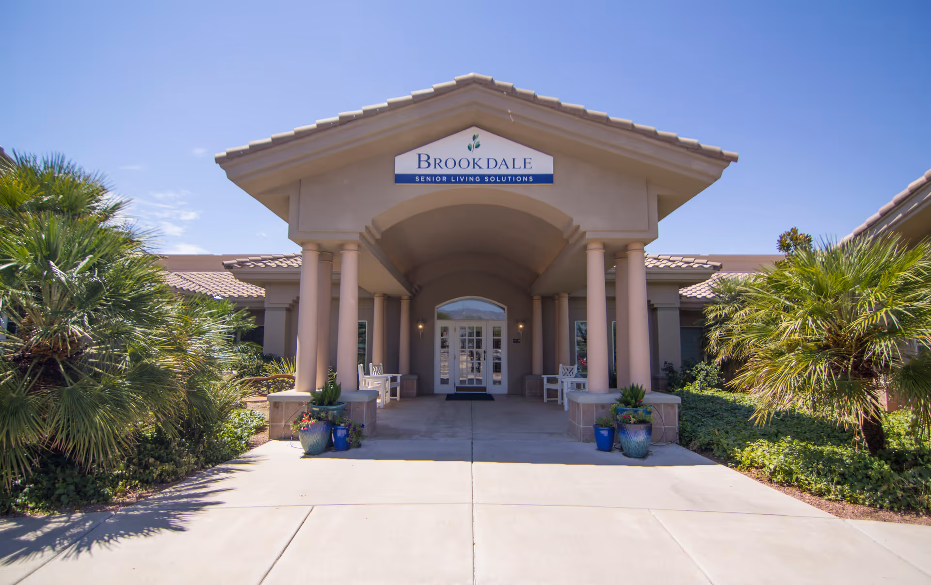 Front entrance of Brookdale Tanque Verde, beige senior living facility with columns and palm trees.