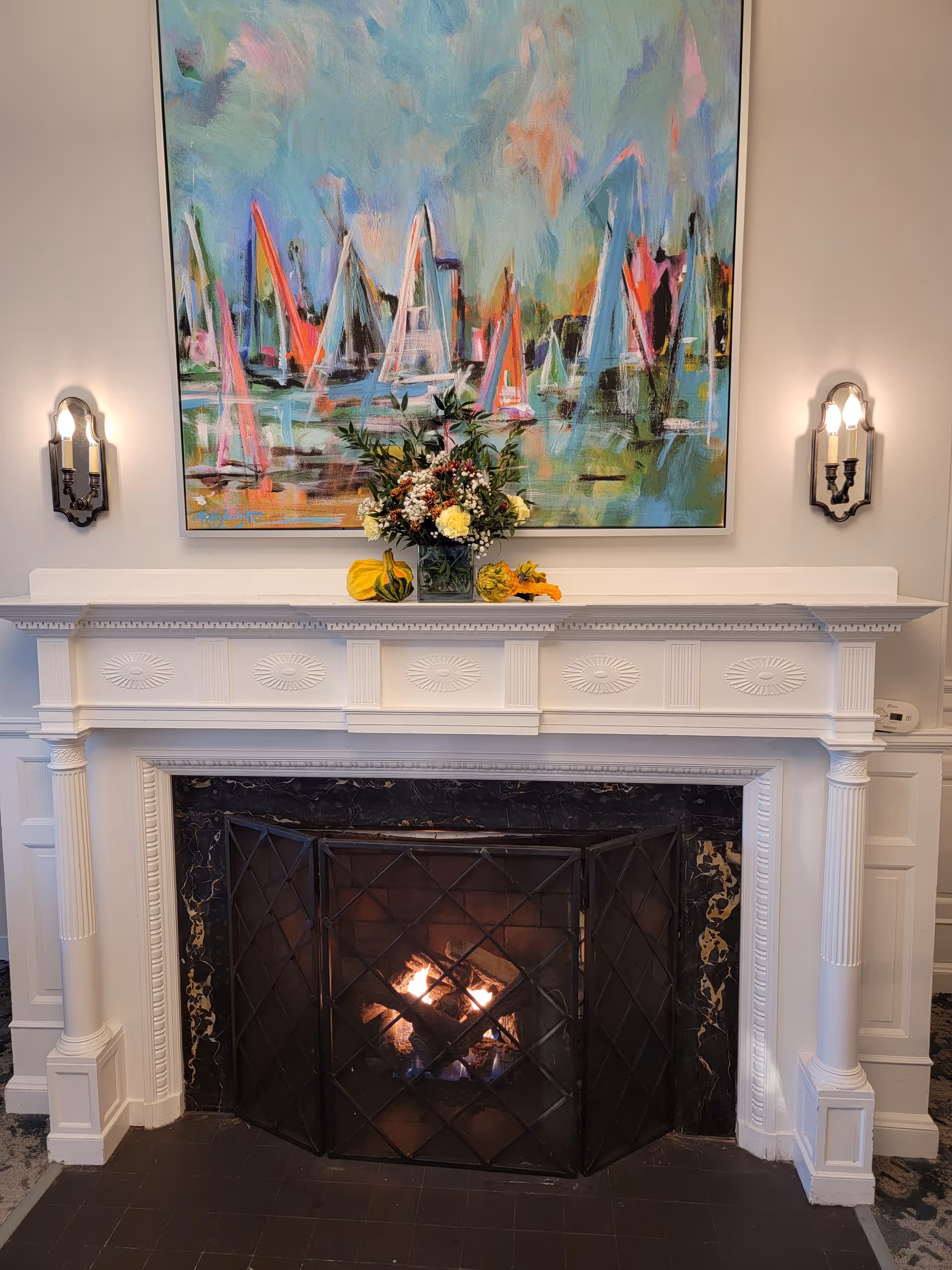 White fireplace with decorative mantel, colorful abstract painting, floral arrangement, and wall sconces.