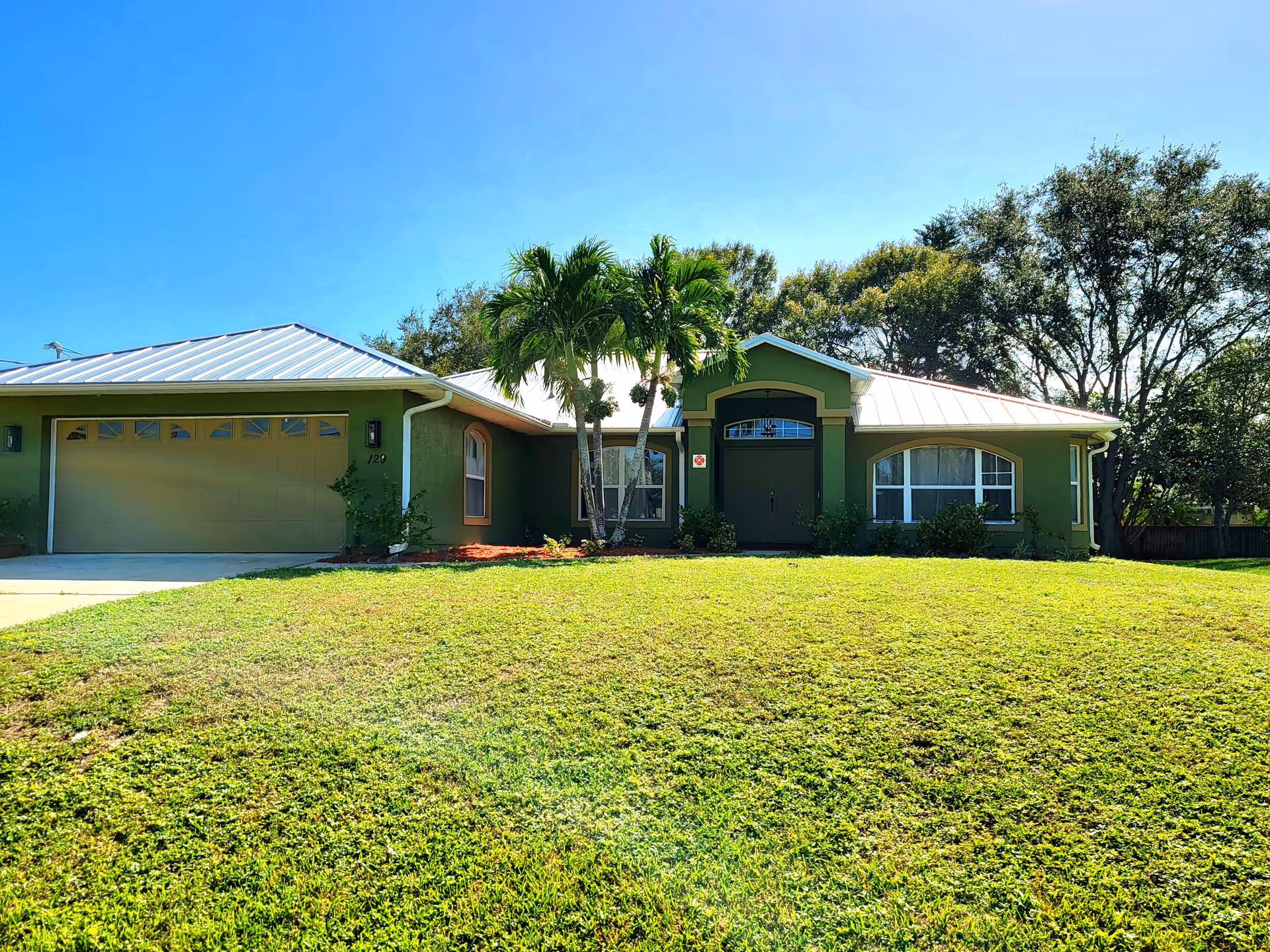 Green single-story building with a two-car garage and palm trees in the front yard