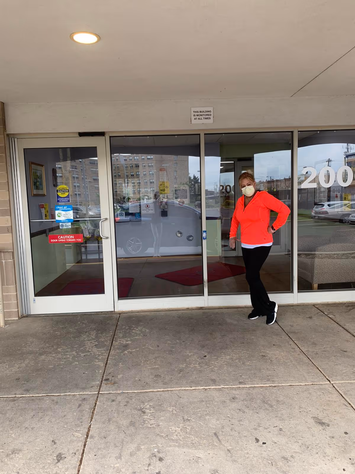 Person wearing a mask stands outside glass doors at a building entrance.