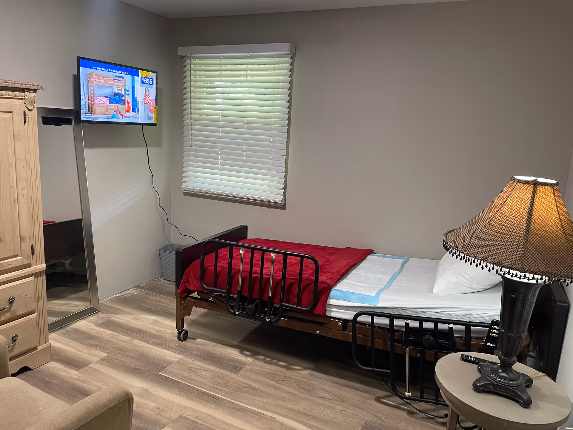 A compact resident bedroom with metal bed frame, dresser, window blinds, and lamp.