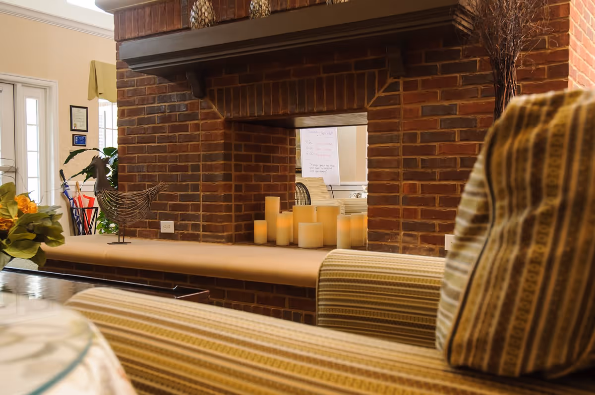 Cozy interior view of a senior living facility featuring a brick fireplace with several decorative candles on the hearth. In the foreground, there is a striped upholstered chair, and in the background, a door with glass panels and some framed certificates on the wall are visible.