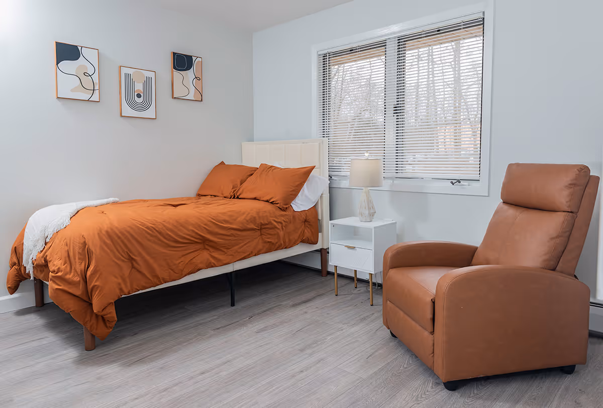 A cozy bedroom with a single bed dressed in rust-colored bedding, a white nightstand with a lamp, a large window with blinds, and a brown leather recliner chair. Three abstract art pieces hang on the wall above the bed.