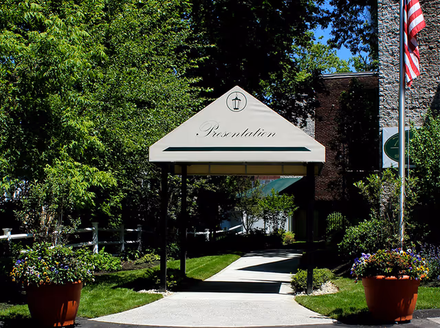 Entrance walkway to Presentation Rehabilitation & Skilled Care Center with a white canopy displaying the word 'Presentation'. The path is flanked by green trees, bushes, and large flower pots. An American flag is visible on the right side near the building.