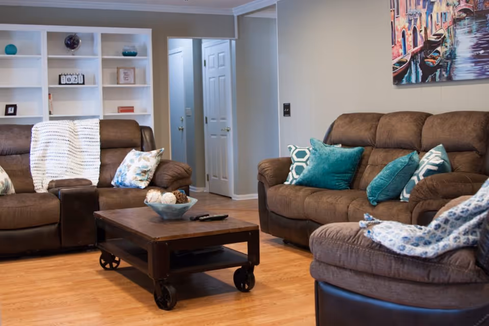 A cozy living room with brown cushioned sofas adorned with decorative pillows and throws. A wooden coffee table with wheels holds a decorative bowl and remote controls. In the background, there is a white built-in bookshelf with various decorative items and a hallway with white doors. A colorful painting hangs on the wall above one of the sofas.