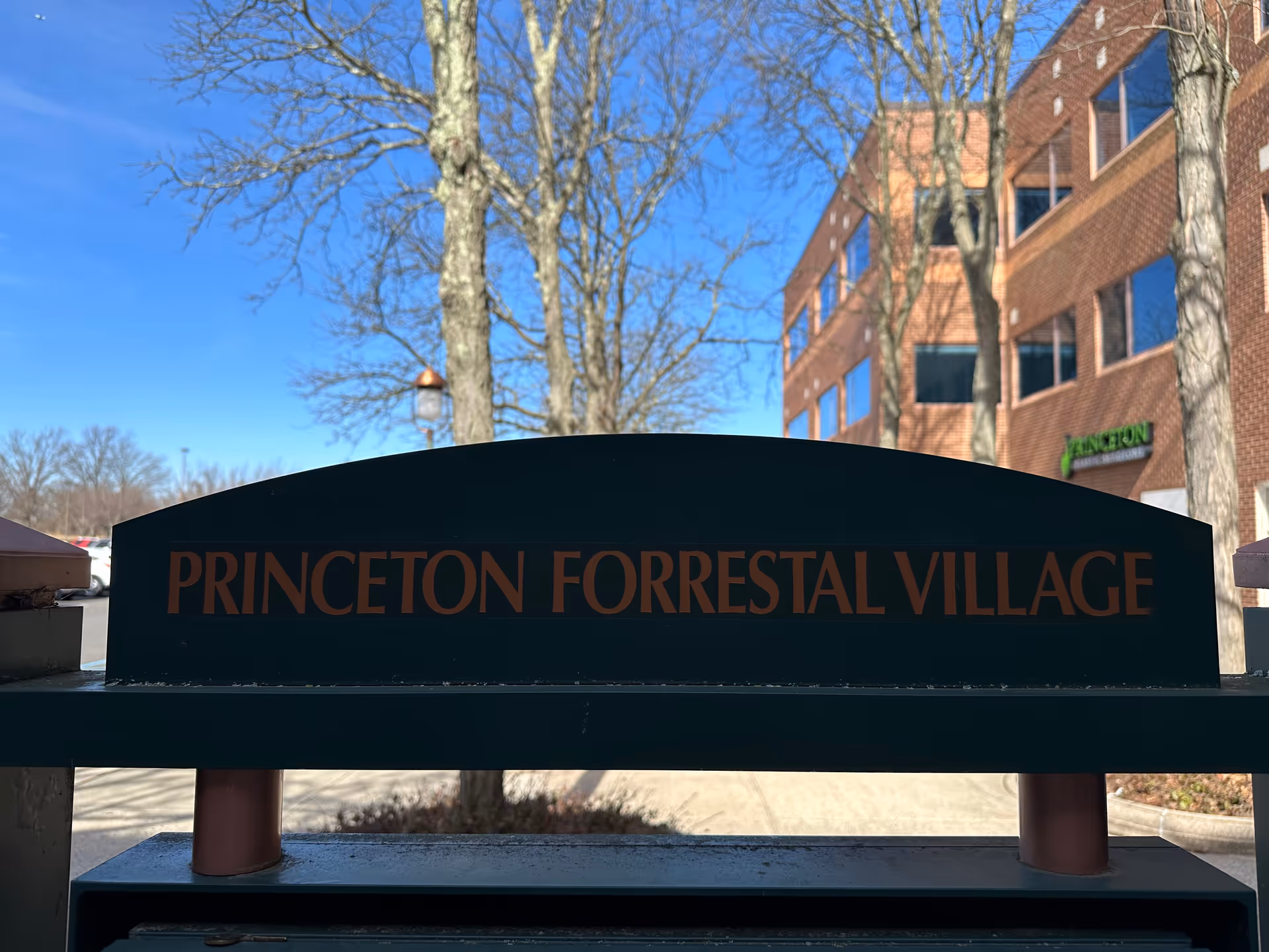 Exterior view of Princeton Forrestal Village sign with brick building and leafless trees.