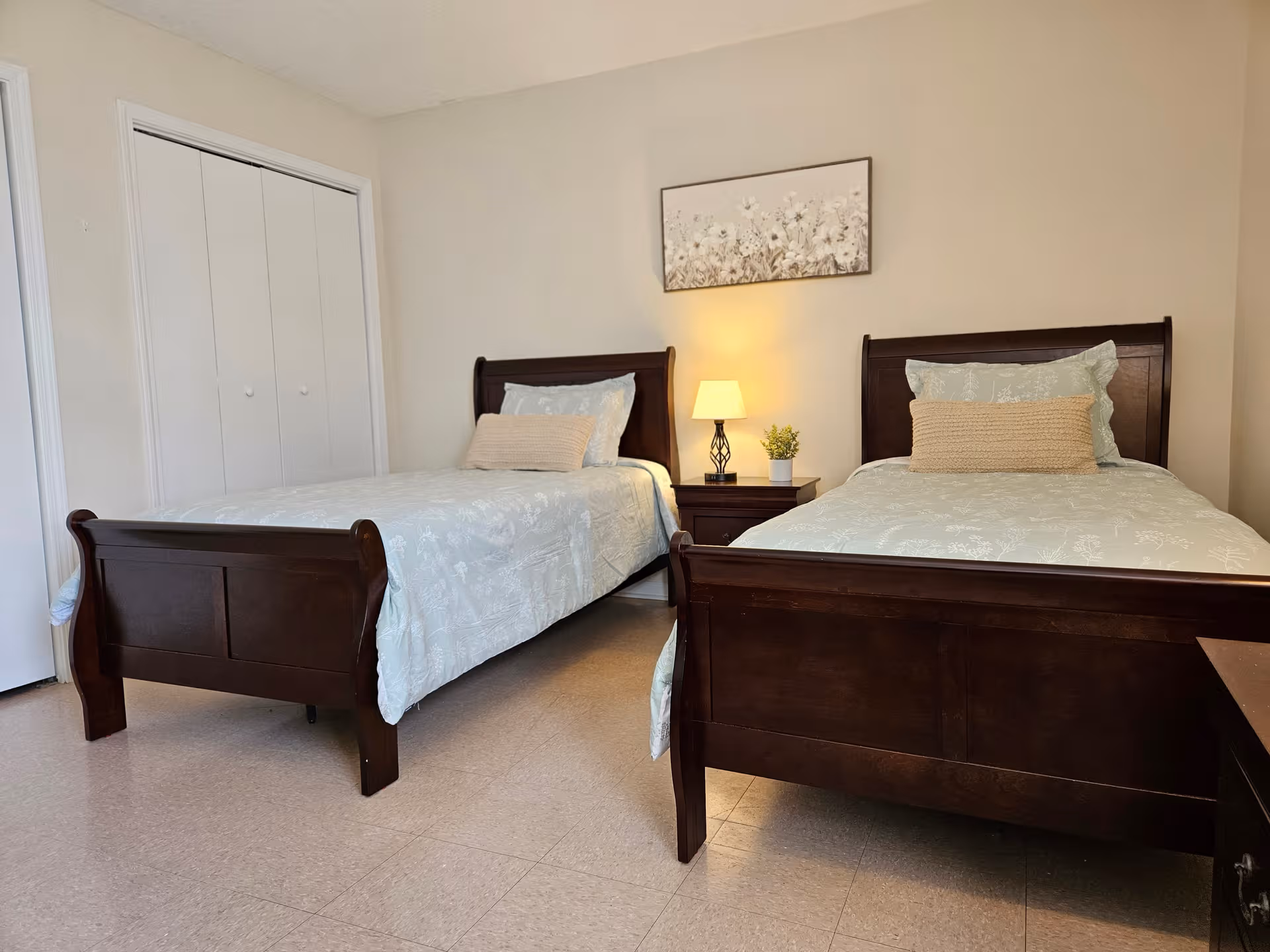 Two dark wood twin beds with bedding, nightstand, lamp, and wall art in a bright room.