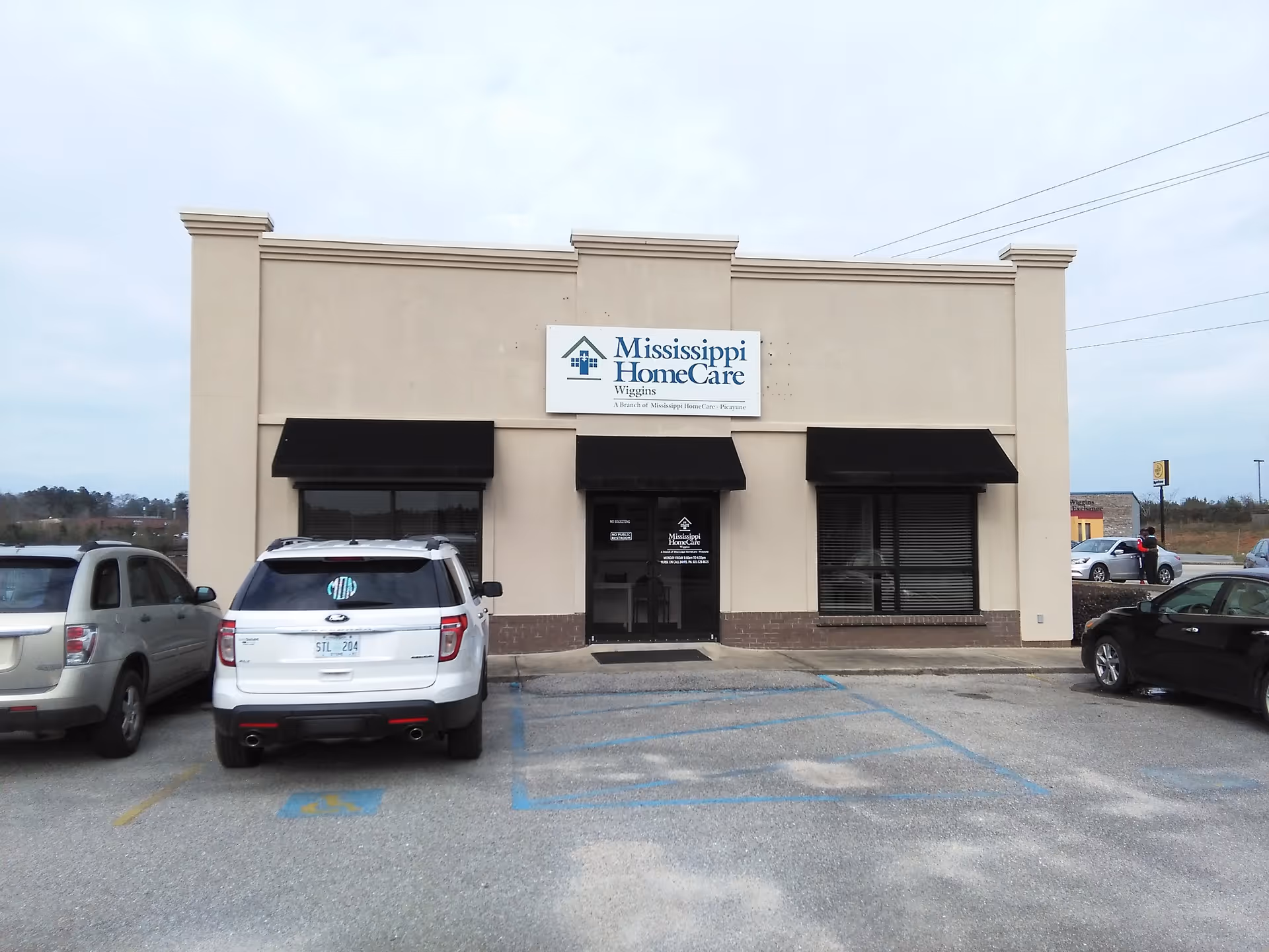 Front exterior of a beige Mississippi HomeCare building with black awnings and parked cars.