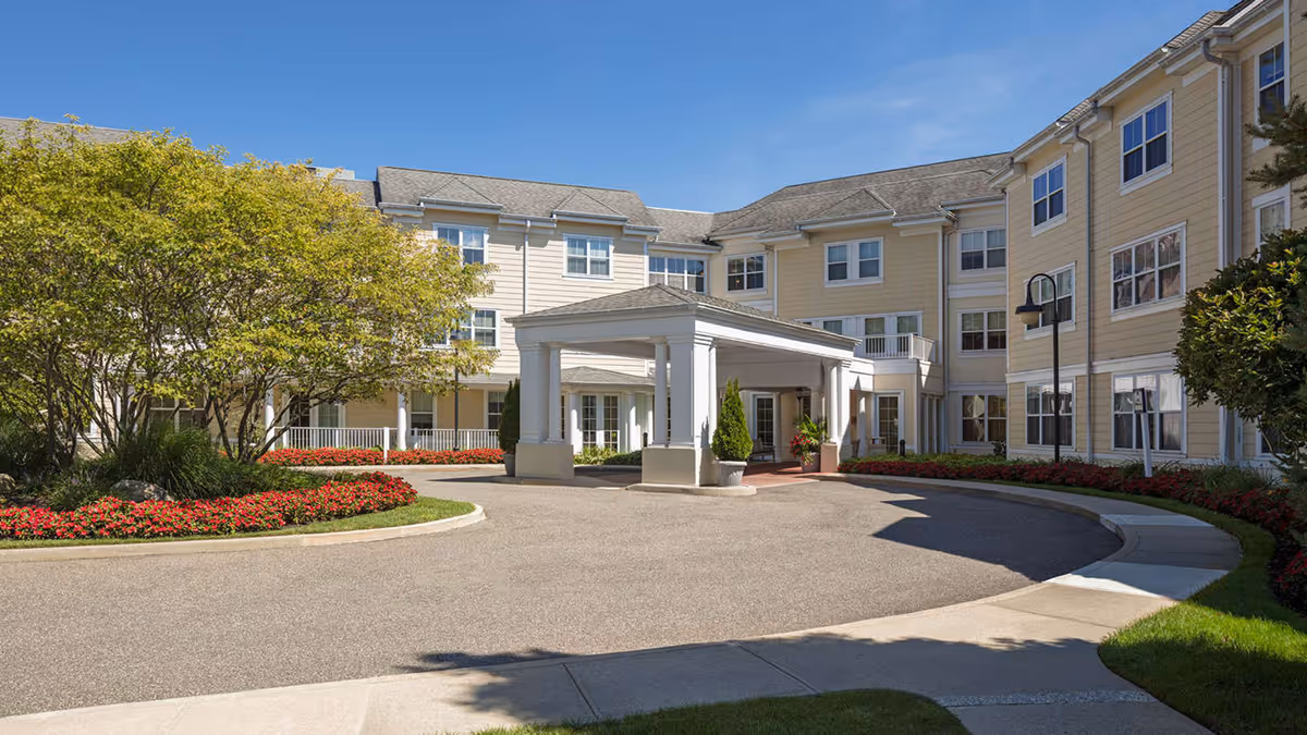 Entrance and circular driveway of a three-story senior living building with a covered porte-cochere and landscaped grounds.