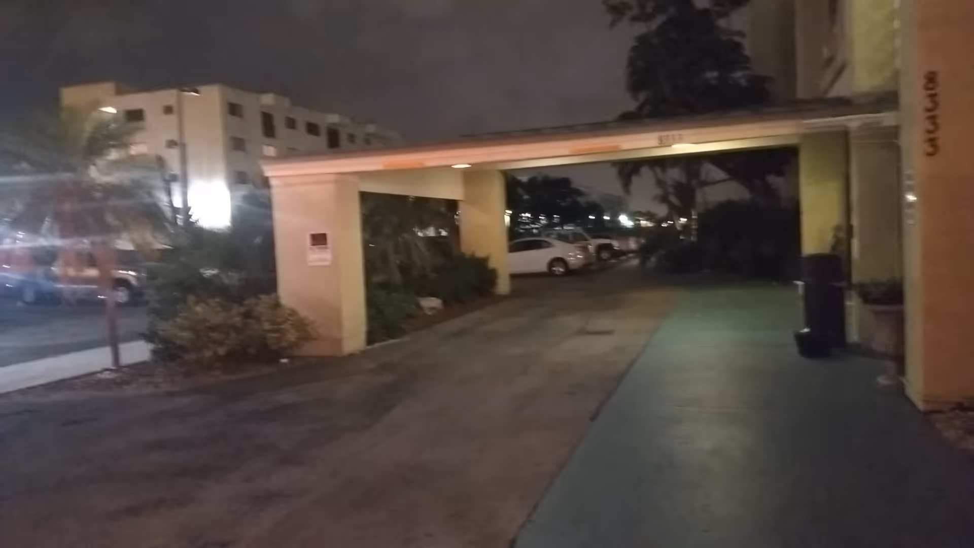 Nighttime exterior of a covered driveway leading to a building entrance with parked cars.