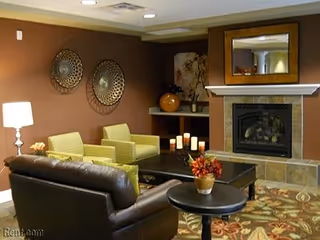 Cozy living room area with a brown leather sofa, two green armchairs, a black coffee table with candles, a small round side table with a flower pot, a fireplace with a large mirror above it, and decorative wall art on a brown wall.