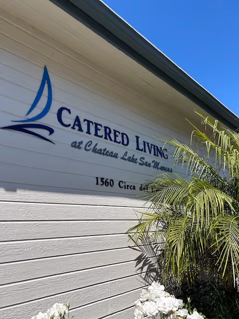 Exterior wall of Chateau Lake San Marcos with 'Catered Living' sign and tropical plant