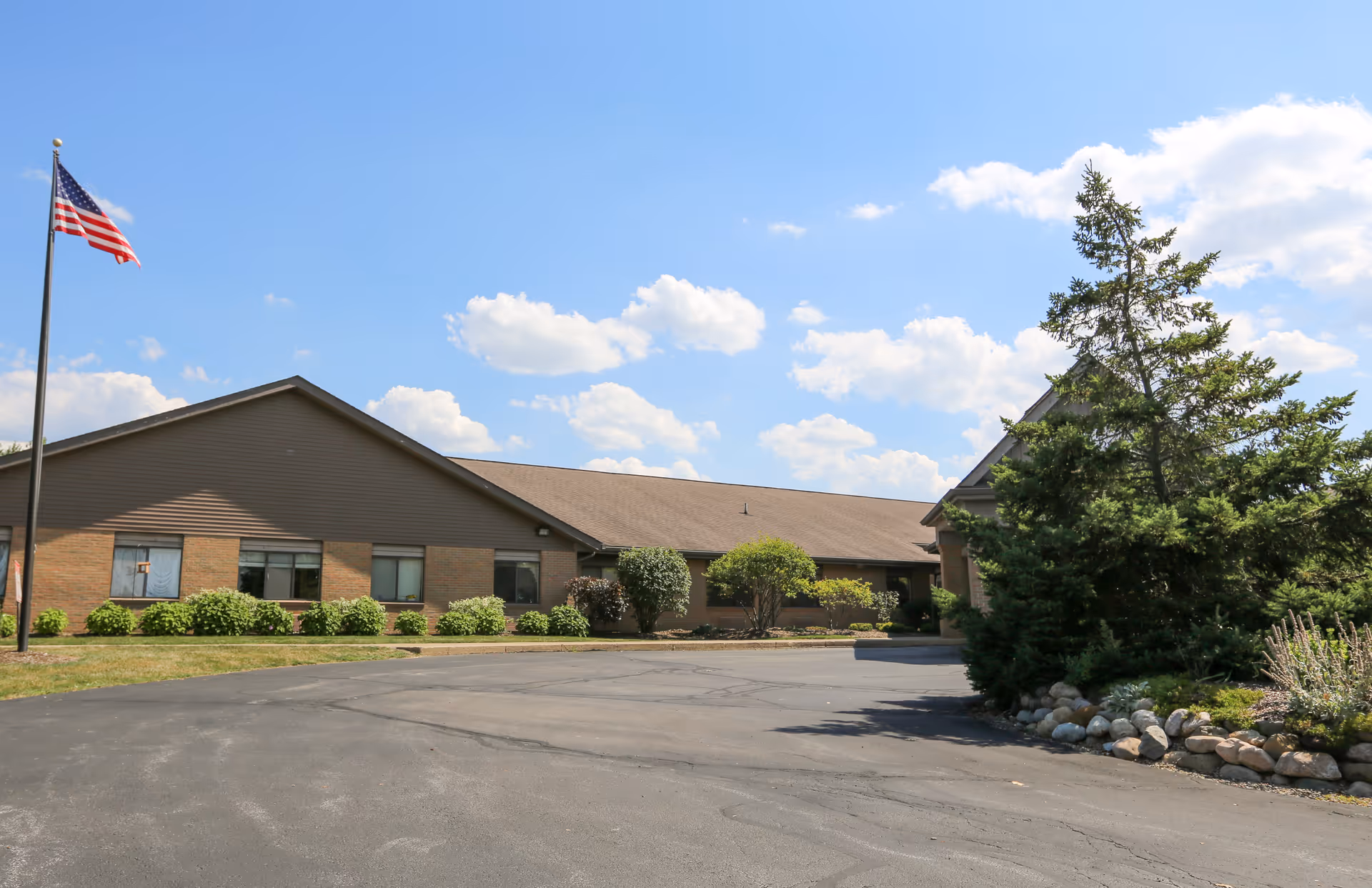 Single-story brick senior living facility with flagpole, manicured shrubs, and a paved driveway.