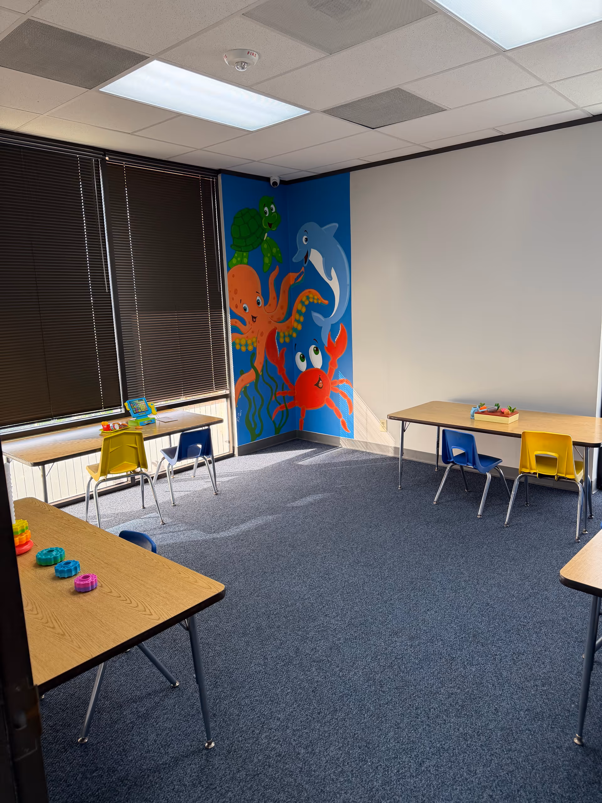 Bright interior children's activity room with tables, chairs, and a mural of sea creatures.