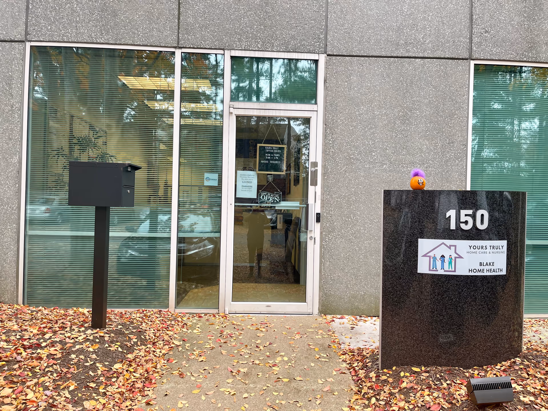 Exterior entrance of a building with glass doors, a tall sign, and fallen autumn leaves.