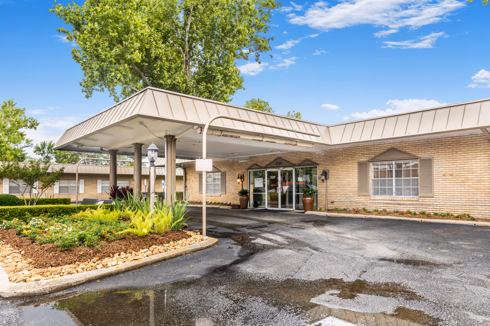 Exterior view of Memorial City Nursing and Rehabilitation Center showing the entrance with a covered drop-off area, brick walls, large windows with shutters, landscaped flower beds, and a clear blue sky.