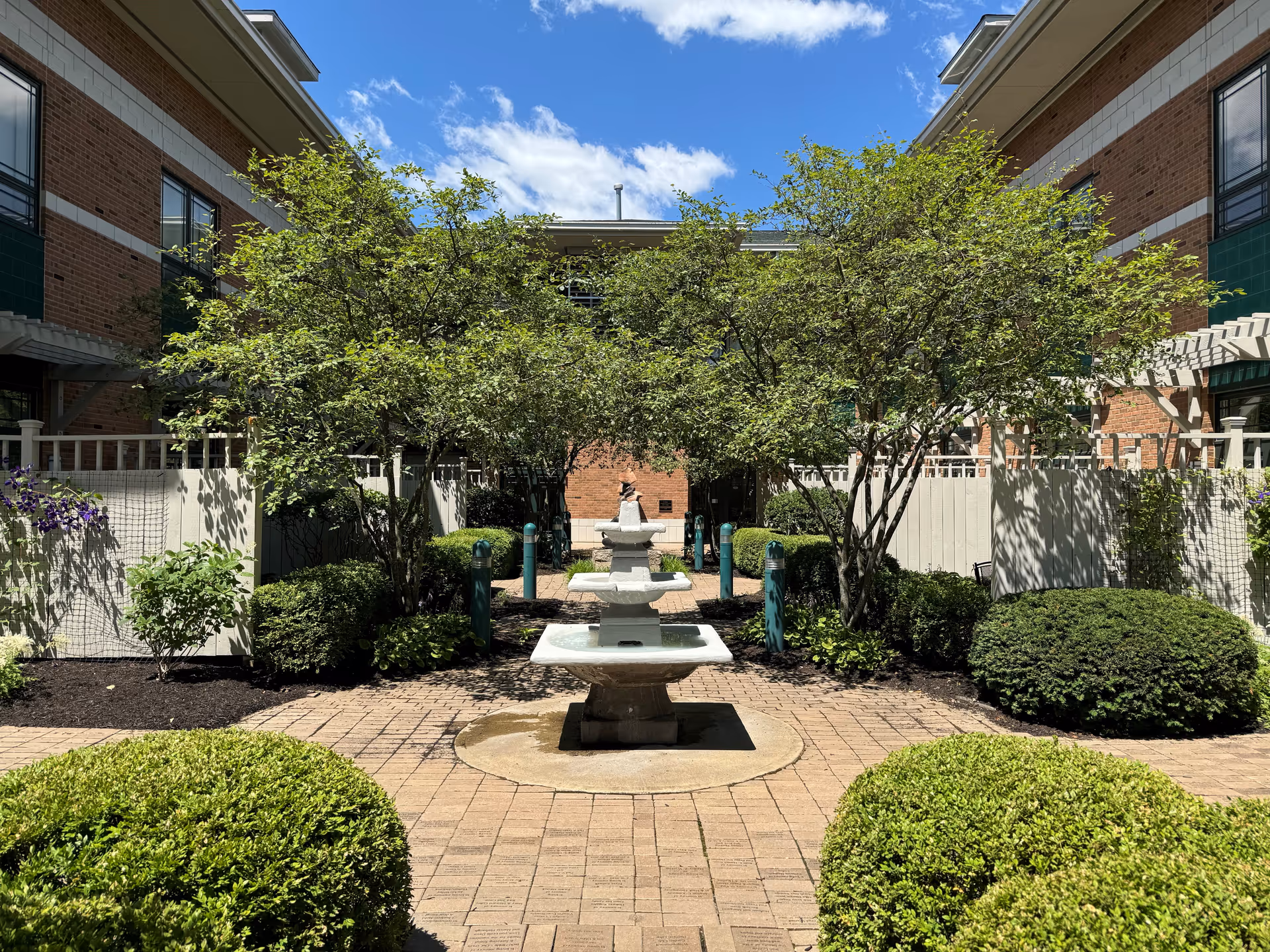 Brick building courtyard with a central multi-tier fountain, trees, and manicured shrubs.