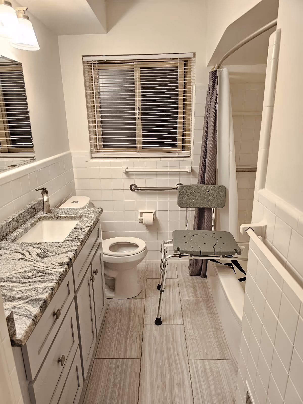 Accessible bathroom with a sink and granite countertop, toilet, shower transfer bench, and grab bars under a window.