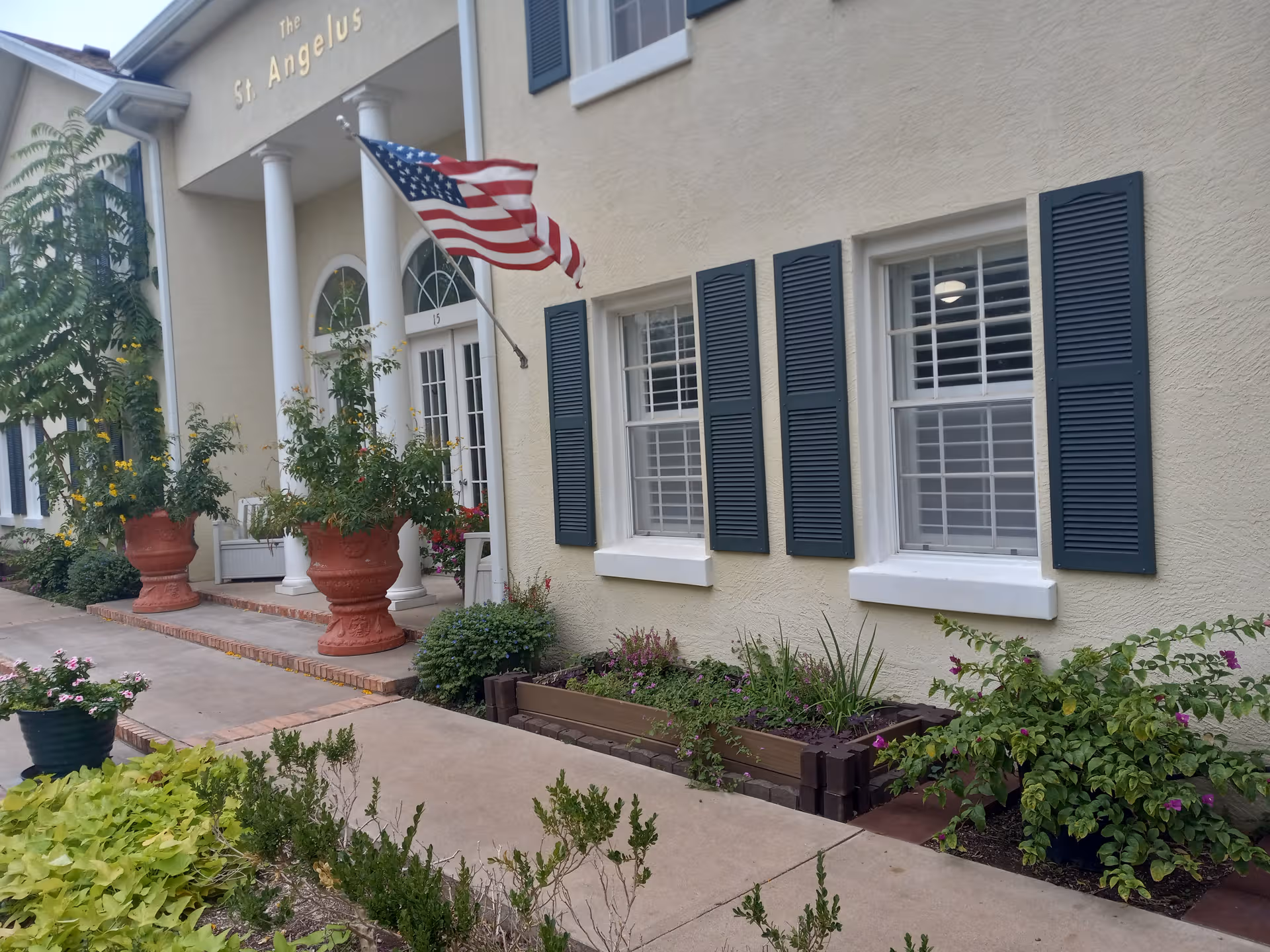 The St. Angelus facility exterior with columns, an American flag, shutters, and flower-filled planters