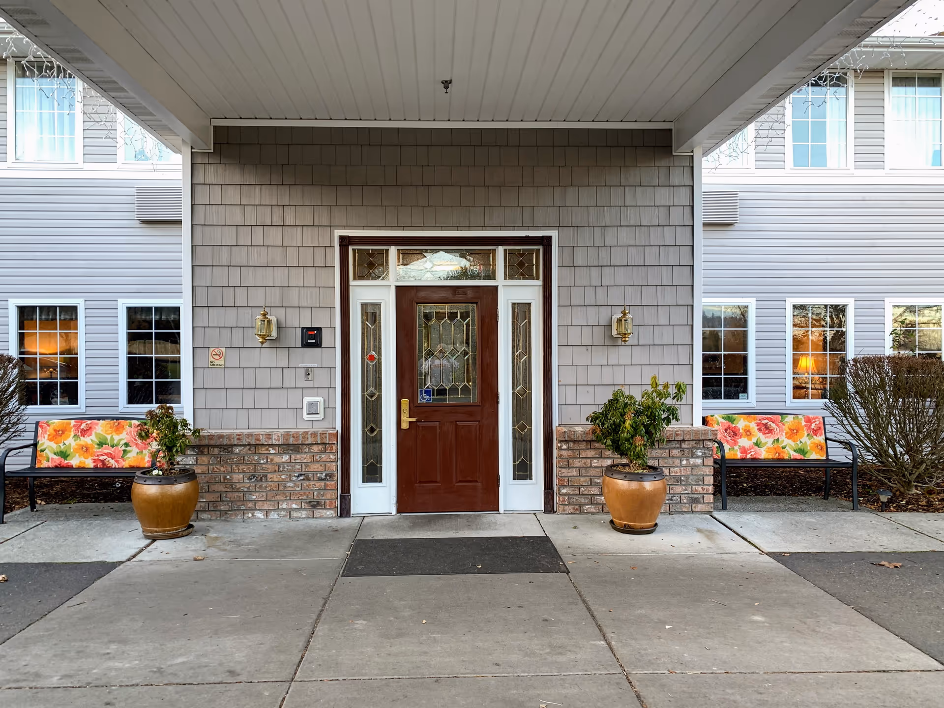 Front entrance of a senior living building with a brick base, glass door, and potted plants.