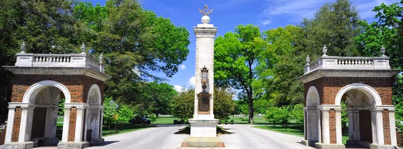 Two brick archways flank a central monument with a tall obelisk in a green park.