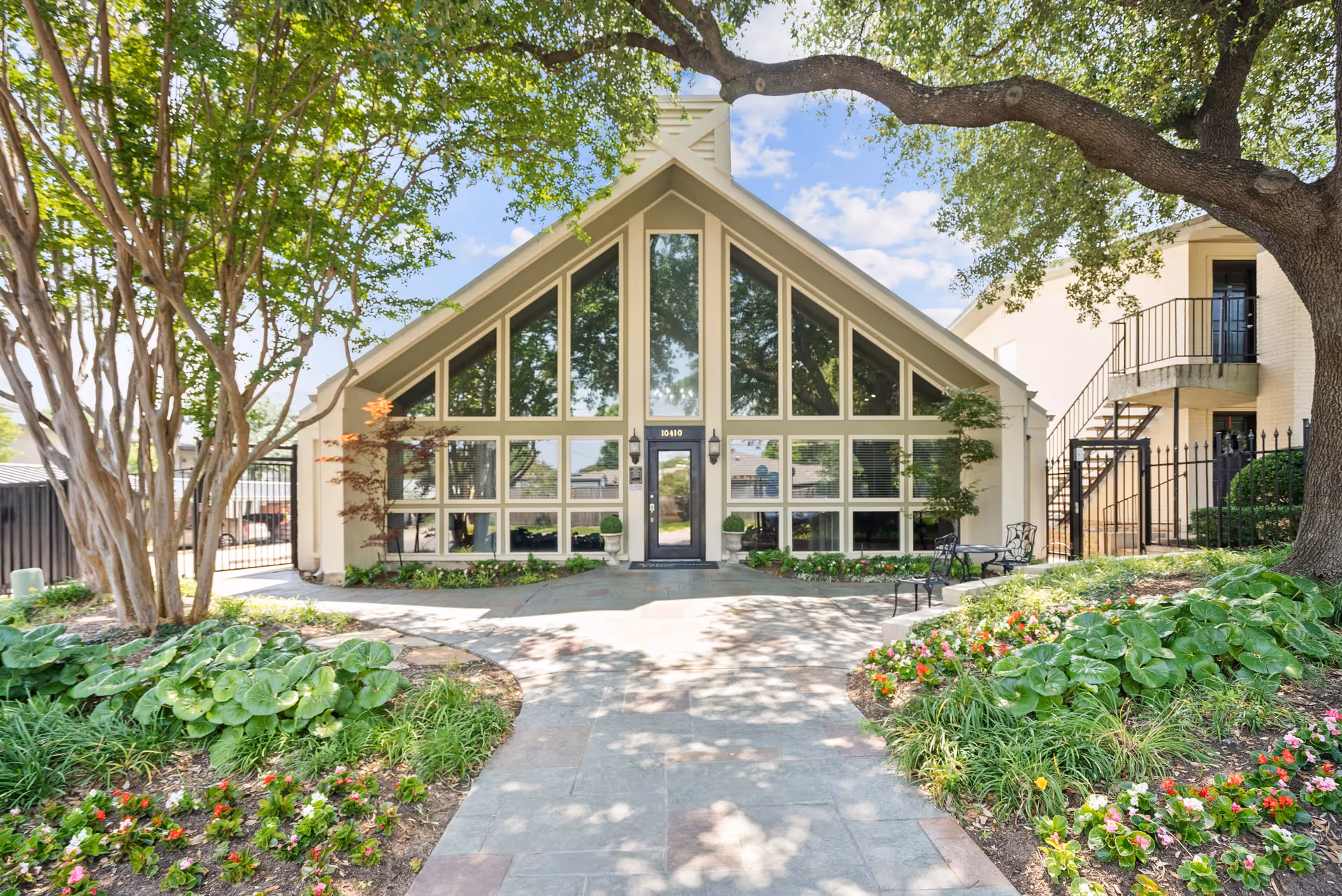 Front exterior view of a building with a large triangular glass facade surrounded by trees, plants, and flowers. A paved walkway leads to the entrance door, with outdoor seating visible on the right side.