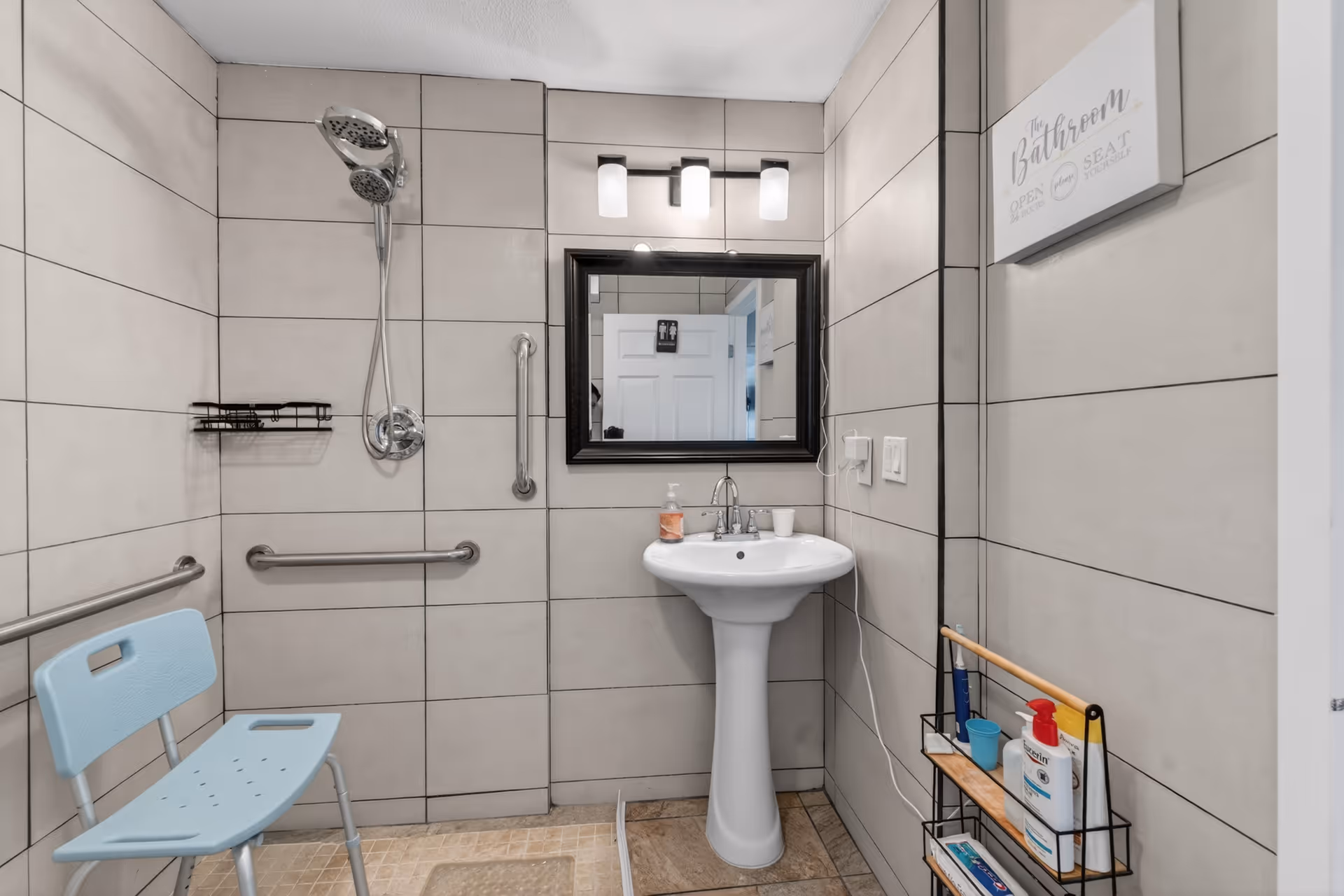 A tiled bathroom with a pedestal sink, mirror, shower with grab bars, and a blue chair.