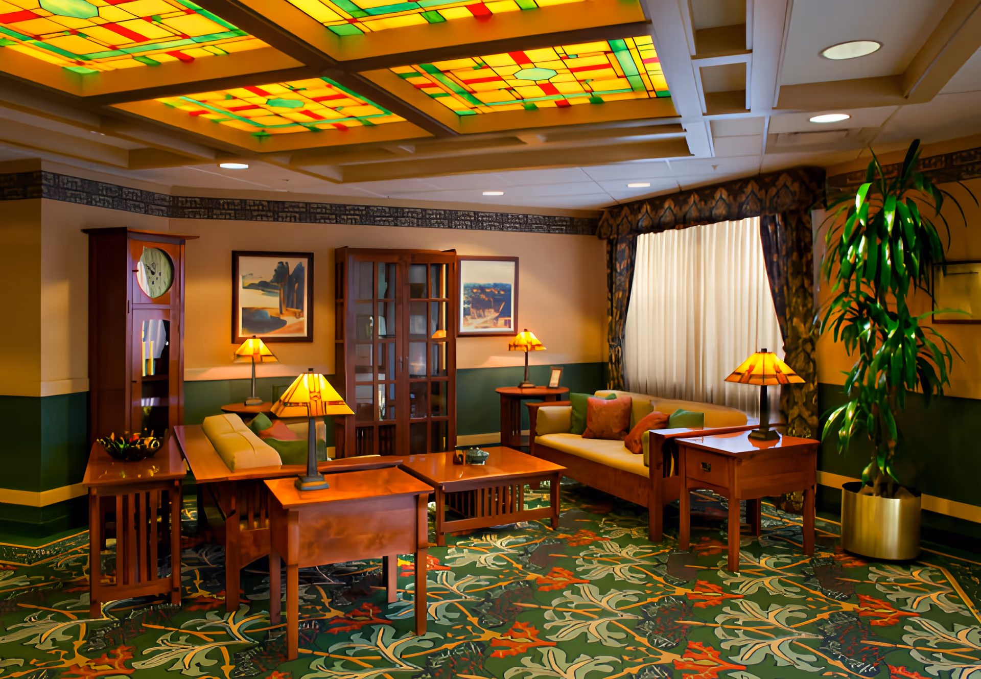 A cozy living room area in a senior living facility featuring wooden furniture including sofas with colorful cushions, side tables with lamps, a tall wooden clock, and a glass-front cabinet. The ceiling has decorative stained glass panels with yellow, green, and red colors. The room has patterned green carpet with floral designs, framed artwork on the walls, a large potted plant, and a window with curtains allowing natural light.