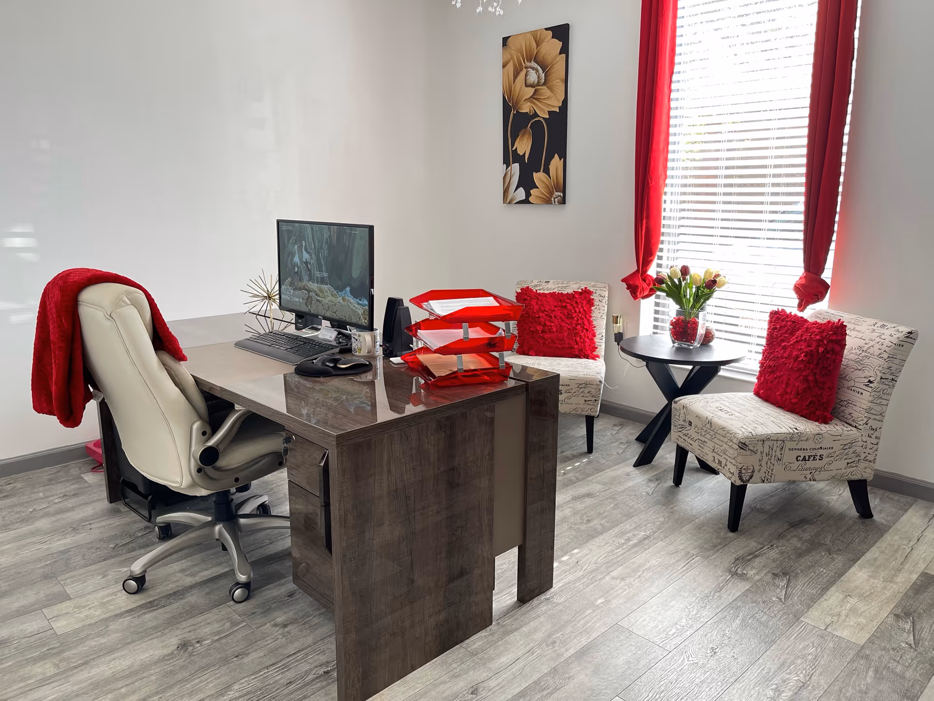 Modern office desk with computer, two patterned chairs, and red cushions near a window.