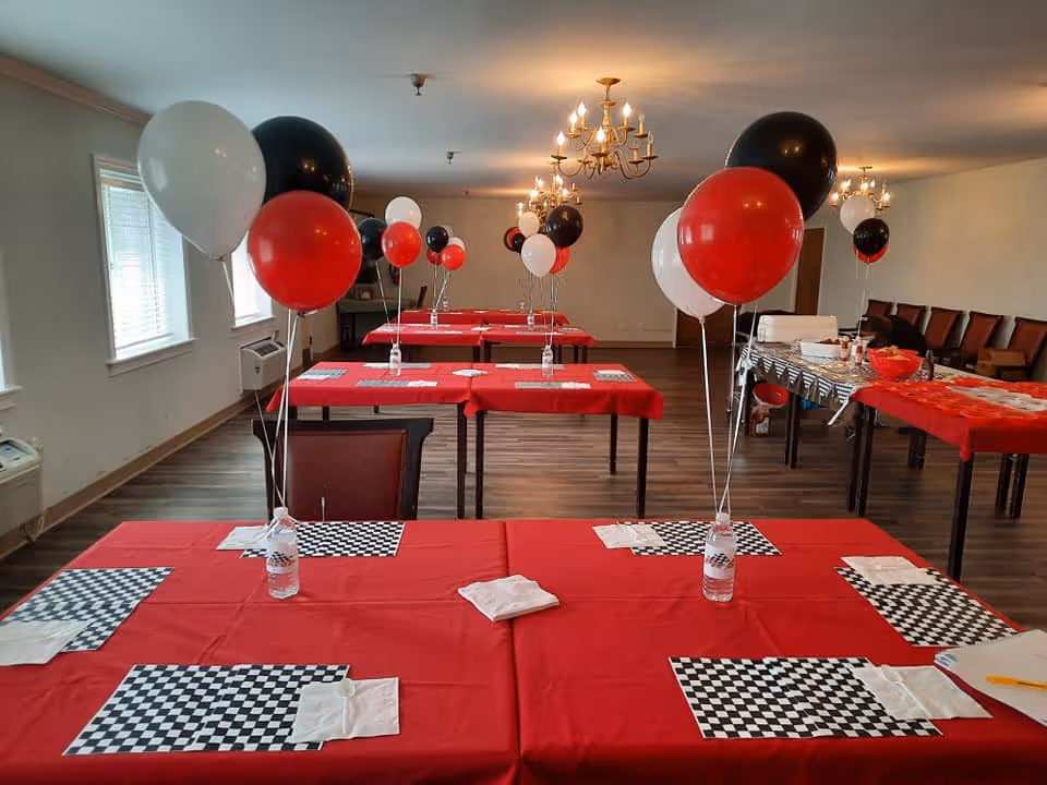 A decorated dining room with tables covered in red tablecloths, each set with black and white checkered placemats, napkins, and water bottles holding red, black, and white balloons. The room has wooden flooring, multiple chandeliers, and chairs lined up against the wall.