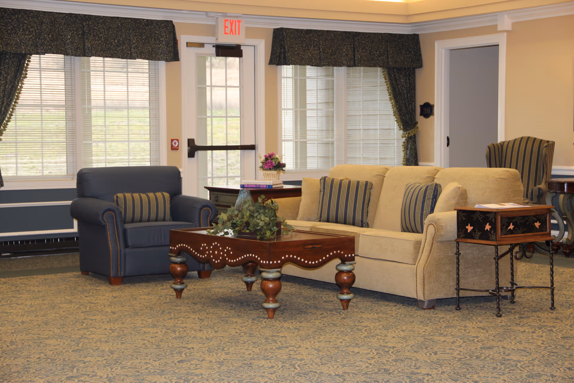 A cozy living room area with a beige sofa adorned with striped cushions, a dark blue armchair with a matching striped pillow, and a wooden coffee table with decorative plants. The room has large windows with blinds and dark green curtains, a patterned carpet, and a side table with a floral arrangement. An exit door is visible in the background.