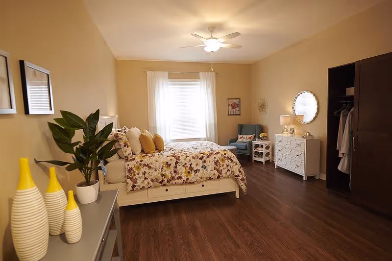 A cozy senior living bedroom with a floral bedspread on a white bed frame, several yellow and white pillows, a blue armchair with a decorative pillow, a white dresser with a round mirror above it, a small white side table with a lamp and flowers, a dark wooden wardrobe with clothes hanging inside, a ceiling fan with light, and a gray table with three decorative yellow and white vases and a potted plant. The room has wooden flooring and beige walls with framed artwork.