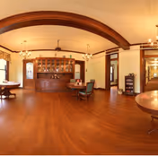 Spacious interior room with polished wooden floors and a large wooden cabinet with glass doors against the wall. Several round tables with chairs are arranged around the room. The ceiling features wooden beams and multiple light fixtures. There are doorways and windows along the walls, and the room has a warm, inviting atmosphere.
