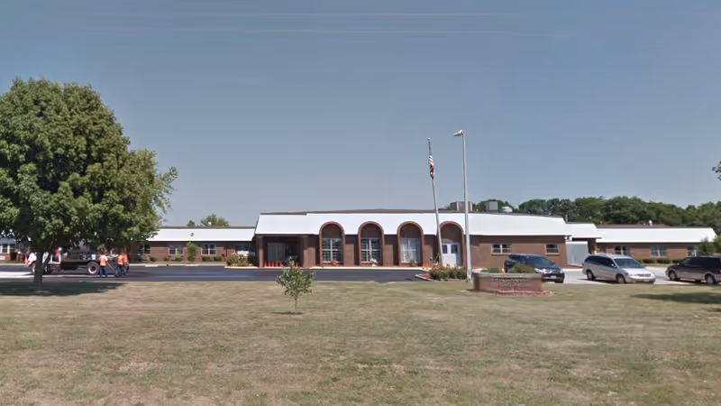 Wide exterior view of a single-story nursing home with arches, flags, and parking.