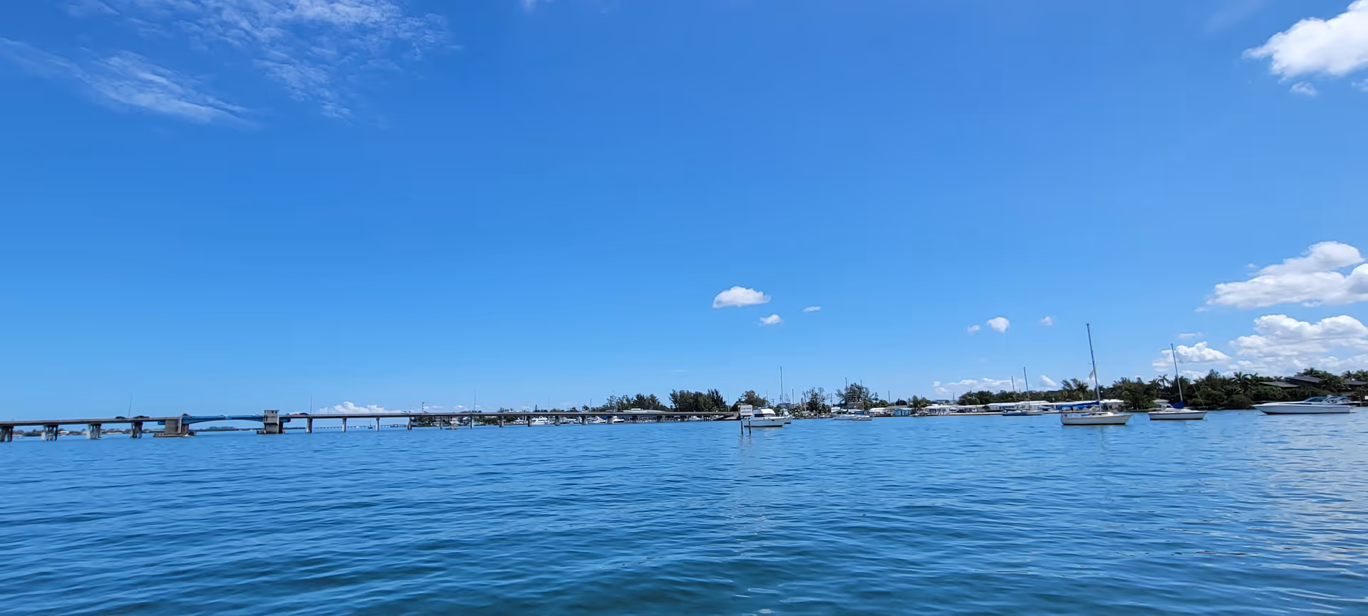 Wide blue water with a distant bridge and boats under a bright sky.
