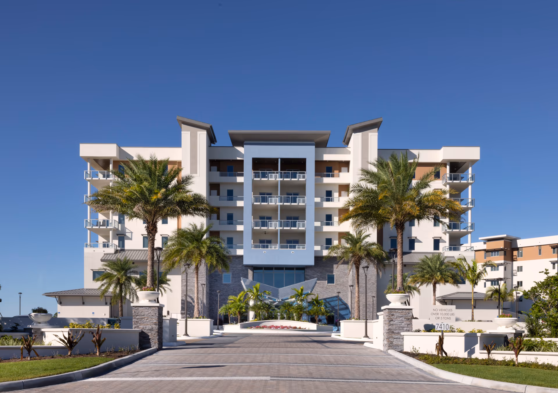 Front exterior view of a modern multi-story senior living facility with balconies, palm trees, and a clear blue sky.
