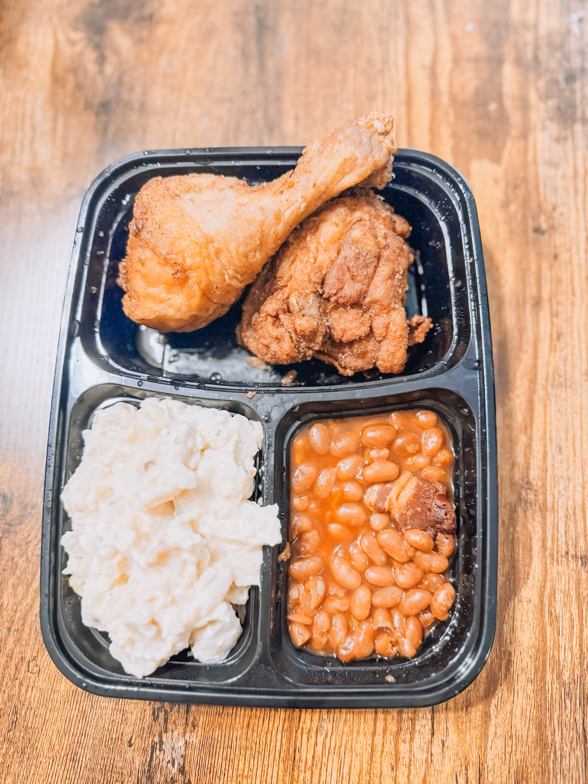 A divided plastic meal tray on a wooden table with fried chicken, mashed potatoes, and beans.
