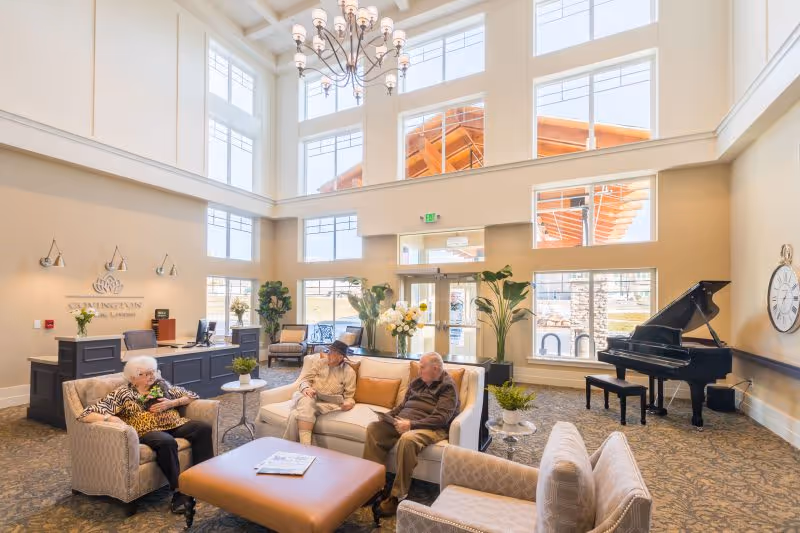 Bright senior living lobby with seating, reception desk, plants, and a grand piano.