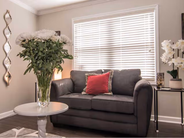 A cozy living room area with a dark gray loveseat featuring a red pillow with a fur trim. In front of the loveseat is a round white table with a large glass vase holding white flowers. To the right, there is a small black side table with a white orchid plant and a decorative candle holder. The background shows a window with white blinds and a decorative wavy mirror on the left wall.